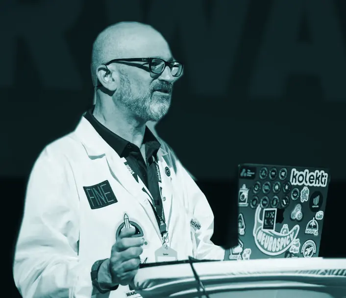 Dr Nick Fine speaking at a conference lectern in glasses and a white lab coat with a FINE patch, a laptop covered in stickers open in front of him